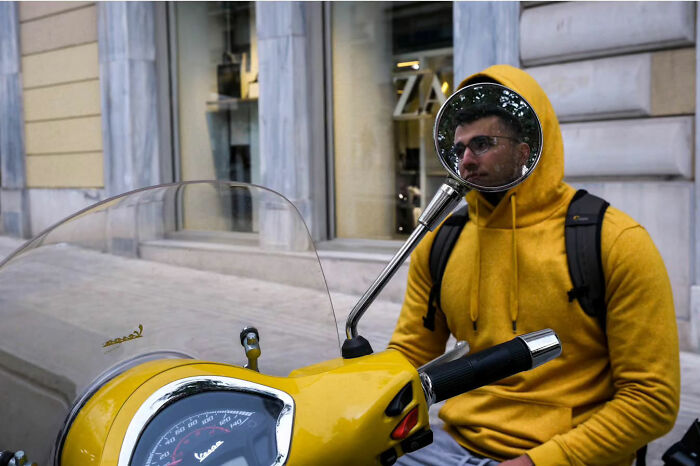 Man in yellow hoodie on a scooter, his face reflected in a mirror, capturing street life's raw beauty.