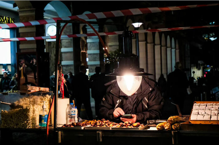 Street life vendor under a lamp, checking his phone while surrounded by popcorn and snacks at a nighttime market.