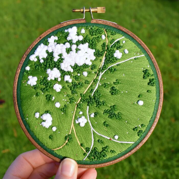 Hand holding intricate landscape embroidery in a hoop, featuring lush green textures and white flowers.