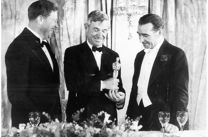 Three men in tuxedos at an awards show, holding an Oscar statuette, with glasses on the table in front.
