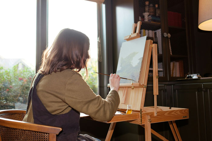 Woman painting on canvas by a window, using a brush to create art in a sunlit room. Woman painting on canvas by a window, using a brush to create art in a sunlit room.