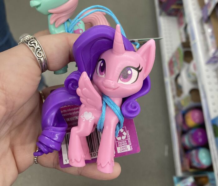 A hand holding a pink unicorn toy with a purple mane in a store; represents whimsical, "useless" items theme.
