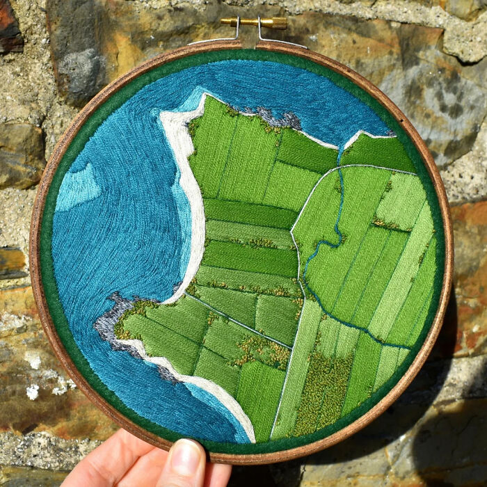 Landscape embroidery by Victoria Rose Richards featuring green fields and a blue ocean, held against a stone wall background.