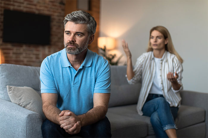 Man in a blue shirt looks pensive while sitting on a couch, woman gestures animatedly beside him. Man in a blue shirt looks pensive while sitting on a couch, woman gestures animatedly beside him.