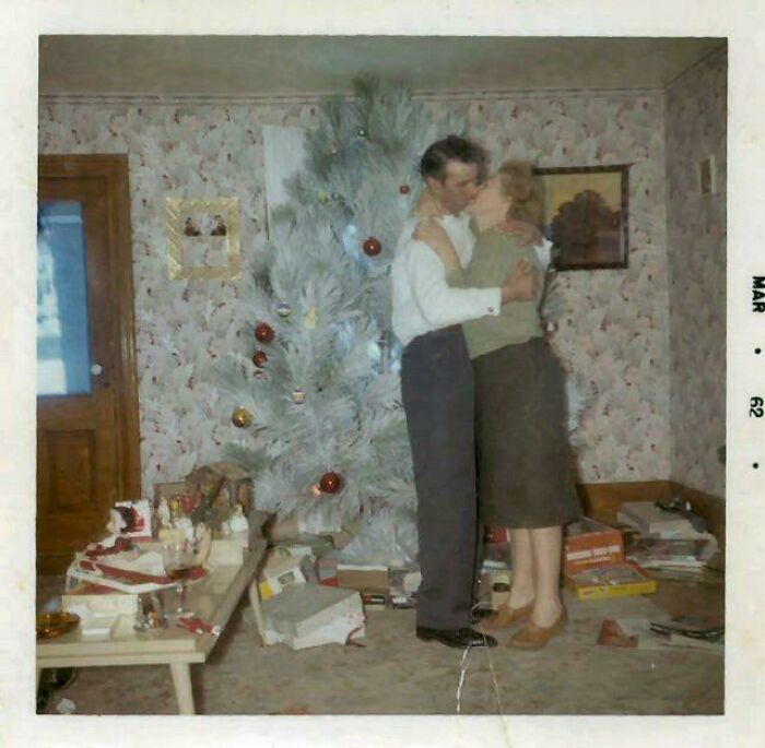 Couple embracing near a white Christmas tree in a 1960s living room, a candid glimpse into the past holiday celebration.