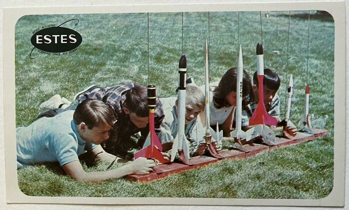 Children lying on grass, setting up vintage model rockets in an outdoor nostalgic hobby scene.