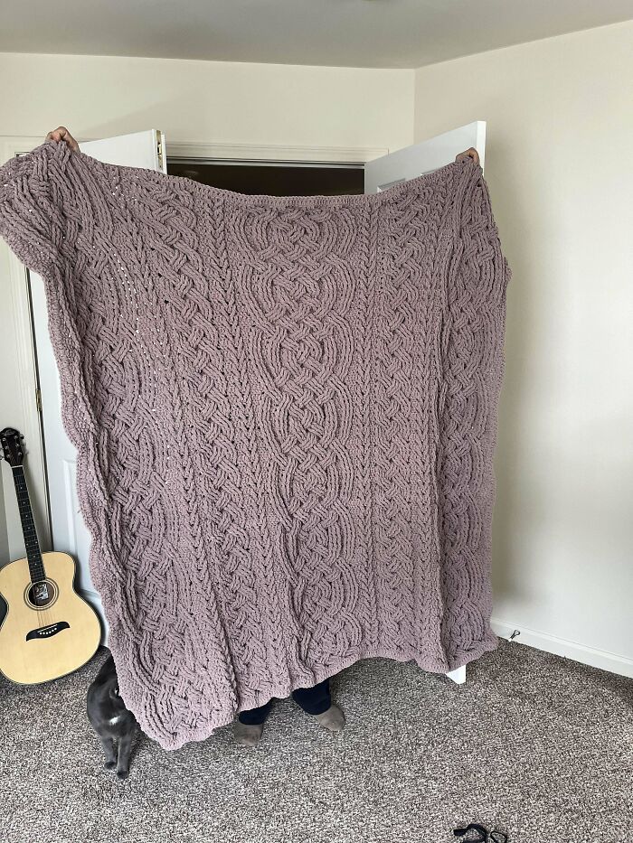 Large hand-crocheted blanket with intricate cable patterns held up in a cozy room, showcasing crochet enthusiasts' incredible work.