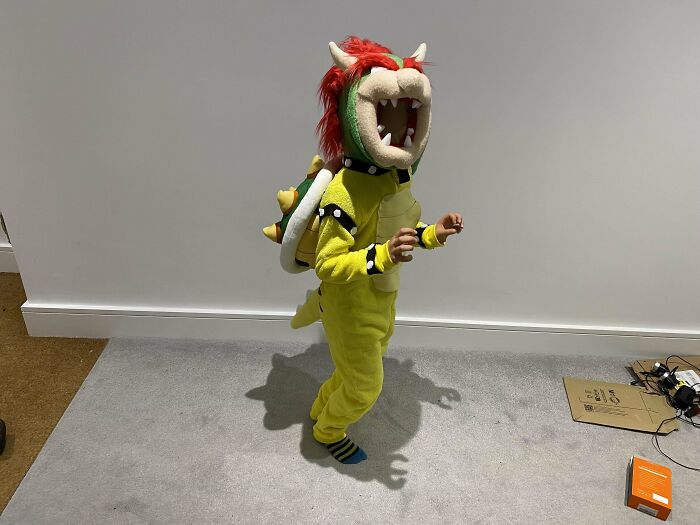 Child in a DIY Bowser costume with a spiky shell and open mouth mask, posing excitedly in a room.