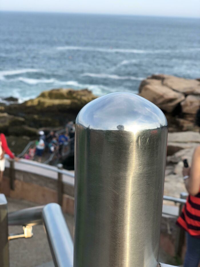 Shiny metal post by the ocean, illustrating things used over time, with rocky background and beach visitors.