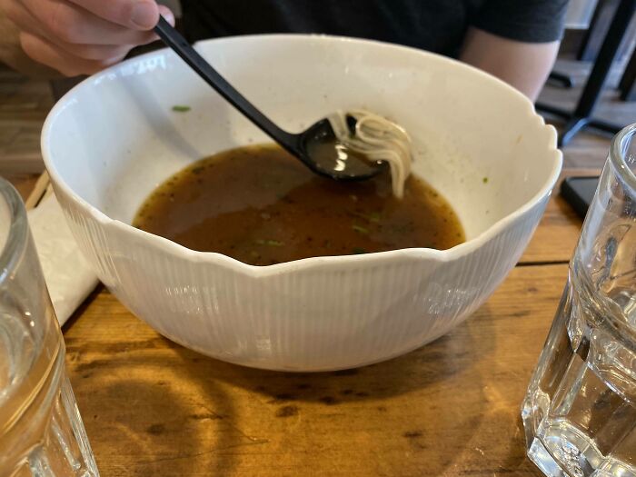 Person holding a spoon with noodles over a large bowl of broth on a wooden table, illustrating minimal things in a meal setting.