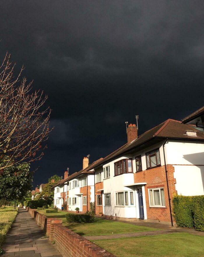 Dark storm clouds loom over a row of houses and a sidewalk, showcasing nature's powerful and terrifying presence.