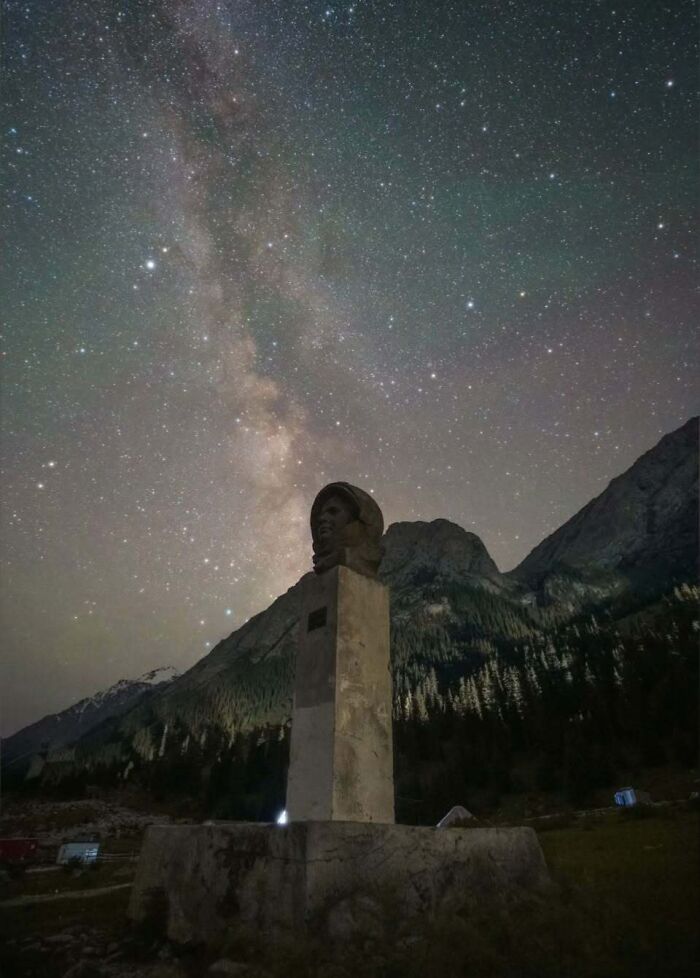 Astrophotography showcases a statue under a starry night sky, with the Milky Way visible against the mountains.