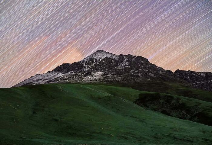 Astrophotography of a mountain under vibrant star trails in the night sky.
