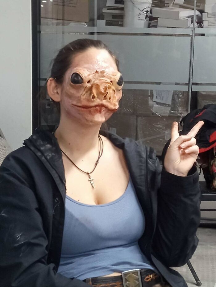 Person wearing a creative DIY alien mask, sitting indoors, giving a peace sign with their hand.