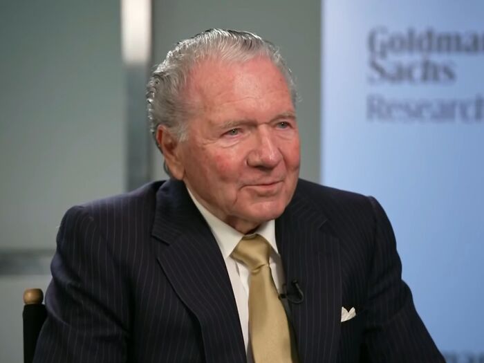 Elderly man in a suit and tie in an interview setting, representing the richest people in the world wealth context.