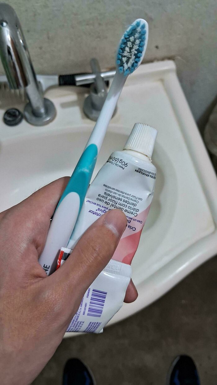 Hand holding a toothbrush and toothpaste tube over a bathroom sink, depicting mundane objects.