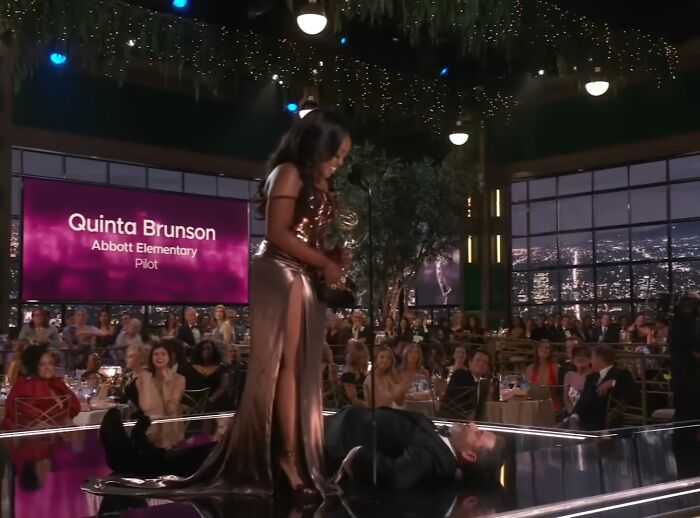 A woman in a gold dress on stage next to a man lying down, during an awards show with audience watching.