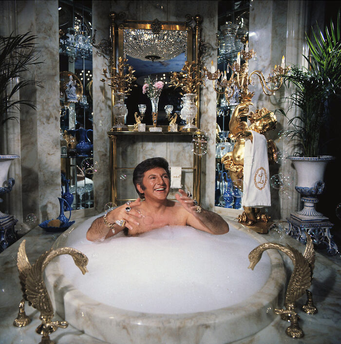 Man in ornate bathtub surrounded by luxurious decor, wearing rings and smiling, capturing a ridiculous moment in retro style.