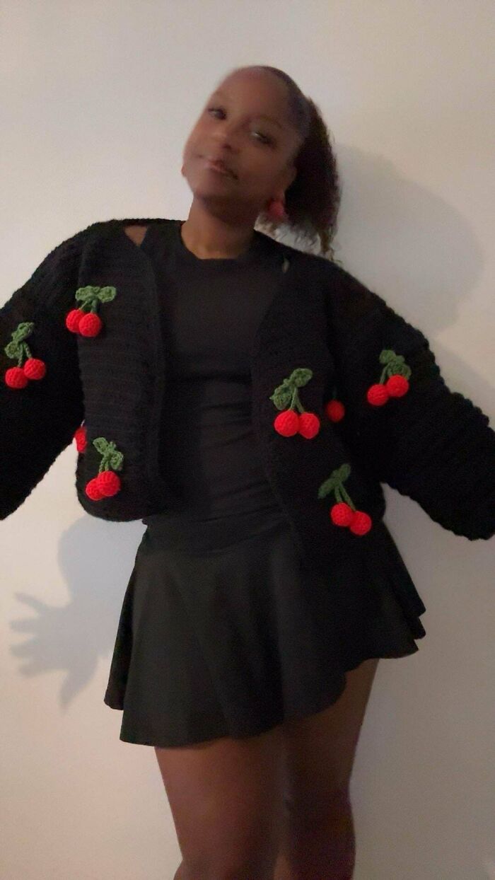 Woman wearing a black crochet cardigan decorated with red cherries, styled with a black top and skirt indoors.