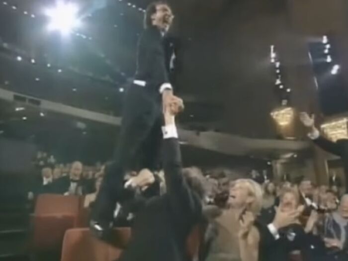 A man balancing on a chair at an awards show, surrounded by a cheering crowd during a scandalous moment.
