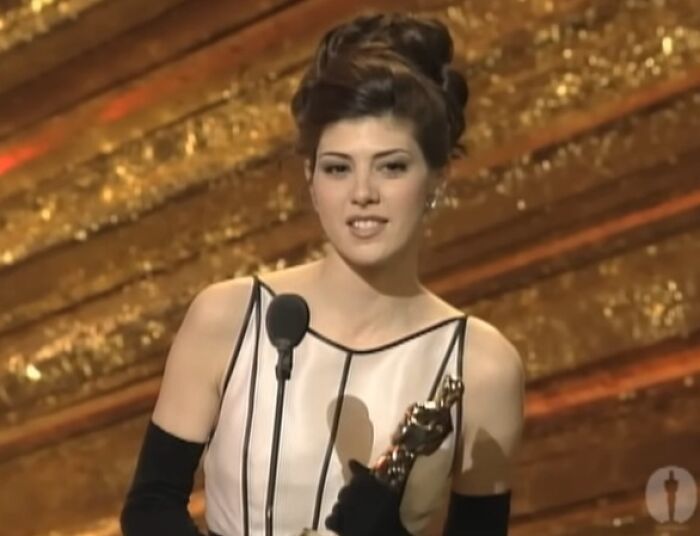Young woman in elegant gown holding award trophy on stage, speaking into a microphone during an awards show.