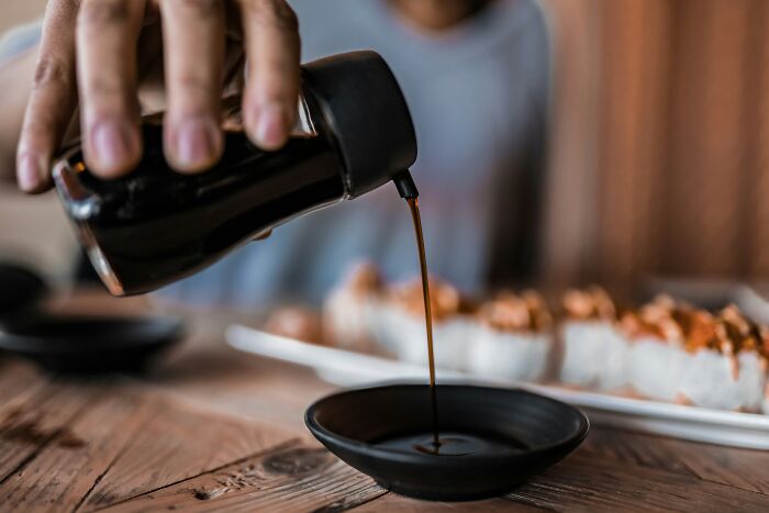 A hand pouring soy sauce into a small dish, with sushi in the background on a wooden table.