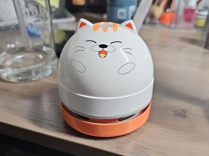 Cute cat-themed desk vacuum, a contender for office Olympics fun, sitting on a wooden desk next to a glass.