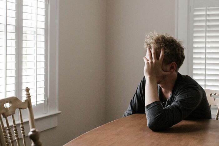 Person sitting at a table with hand on face, frustrated, in a bright room, illustrating stereotypes.