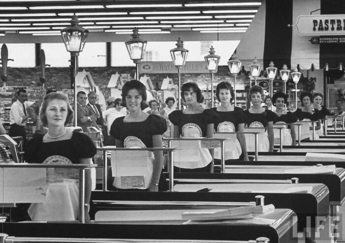 Checkout lines with women in uniform in a 1960s American supermarket, showcasing JFK era America.