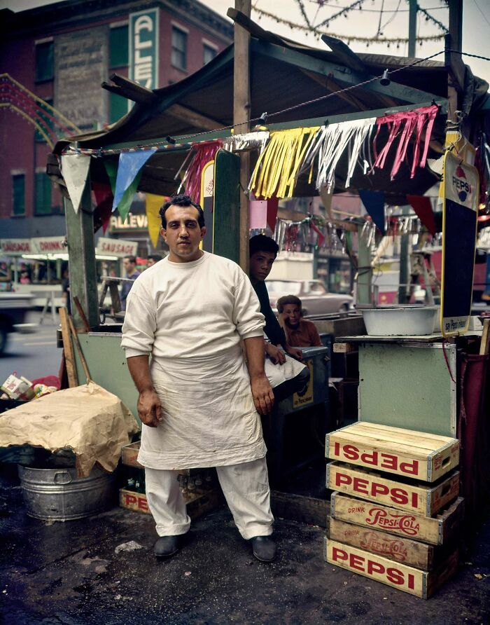 Man in a white apron at a street food stall during the JFK years, surrounded by Pepsi crates and colorful decorations.