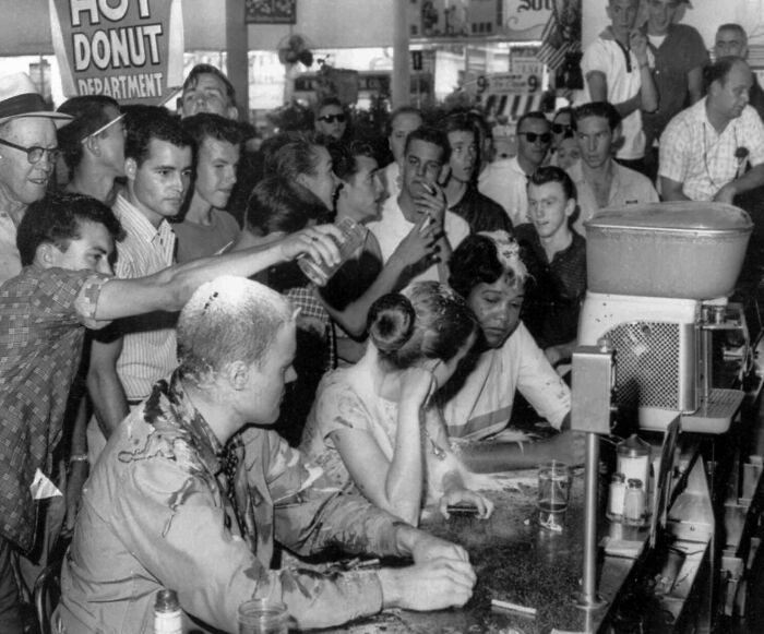 Crowd at a diner counter during the JFK years, with young people gathered, some engaging in a sit-in protest.
