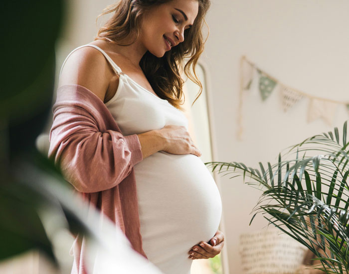 Pregnant woman smiling and holding her belly, wearing a white dress and pink cardigan, next to indoor plants. Pregnant woman smiling and holding her belly, wearing a white dress and pink cardigan, next to indoor plants.