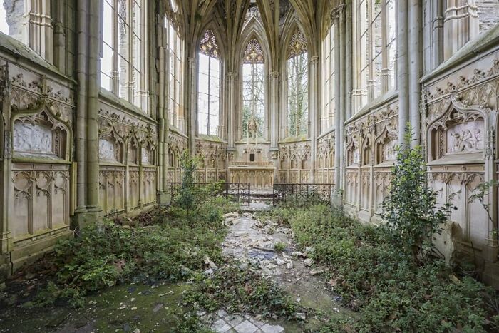 Abandoned, overgrown chapel interior with detailed stone carvings and large windows.
