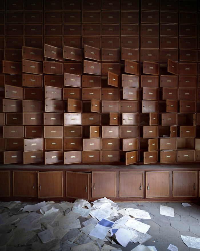 A wall of open drawers in an abandoned location, with scattered papers covering the floor, creating an eerie atmosphere.