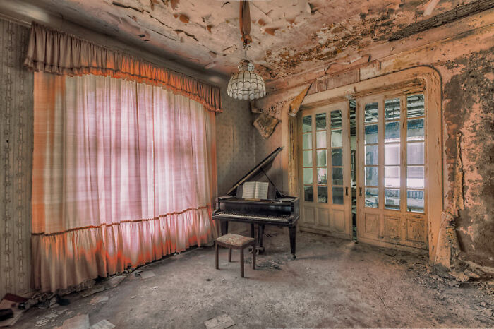 Abandoned place with a dusty piano in a decaying room, featuring peeling walls, a broken window, and tattered curtains.
