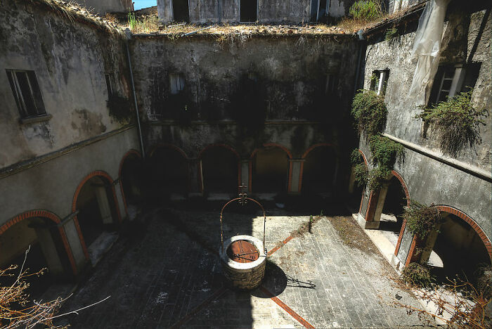 Abandoned courtyard with a central well and overgrown vegetation, featuring eerie, decaying walls and arches.