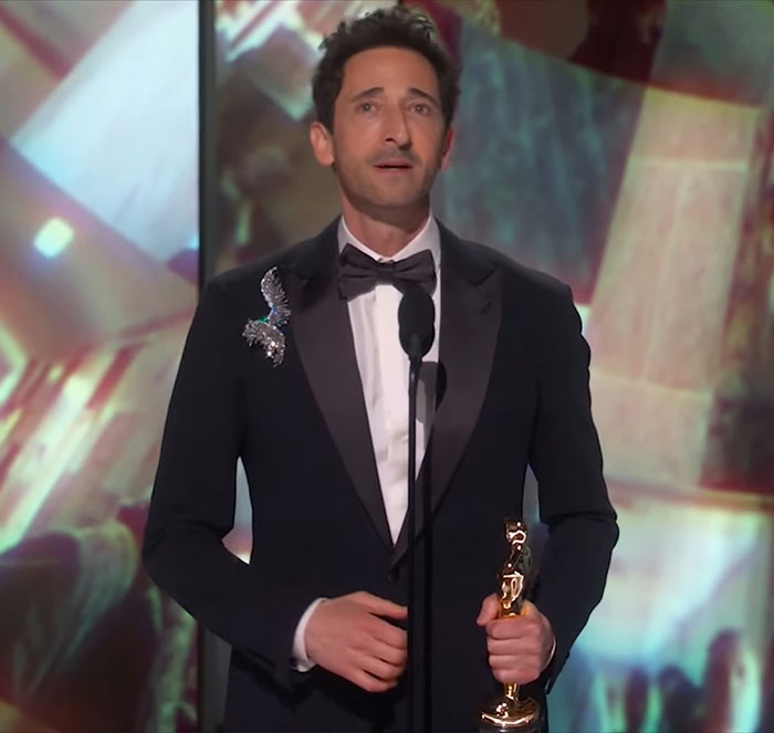 Adrien Brody at the Oscars holding a trophy, wearing a black tuxedo with a butterfly pin. Adrien Brody at the Oscars holding a trophy, wearing a black tuxedo with a butterfly pin.