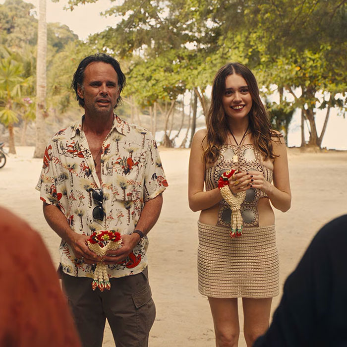 A scene from "The White Lotus" with Aimee Lou Wood, outdoors, holding flowers and smiling by a beach. A scene from "The White Lotus" with Aimee Lou Wood, outdoors, holding flowers and smiling by a beach.