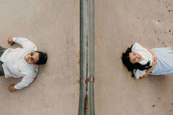 Two people lie on a concrete surface divided by a metal beam, capturing unique engagement photo creativity.