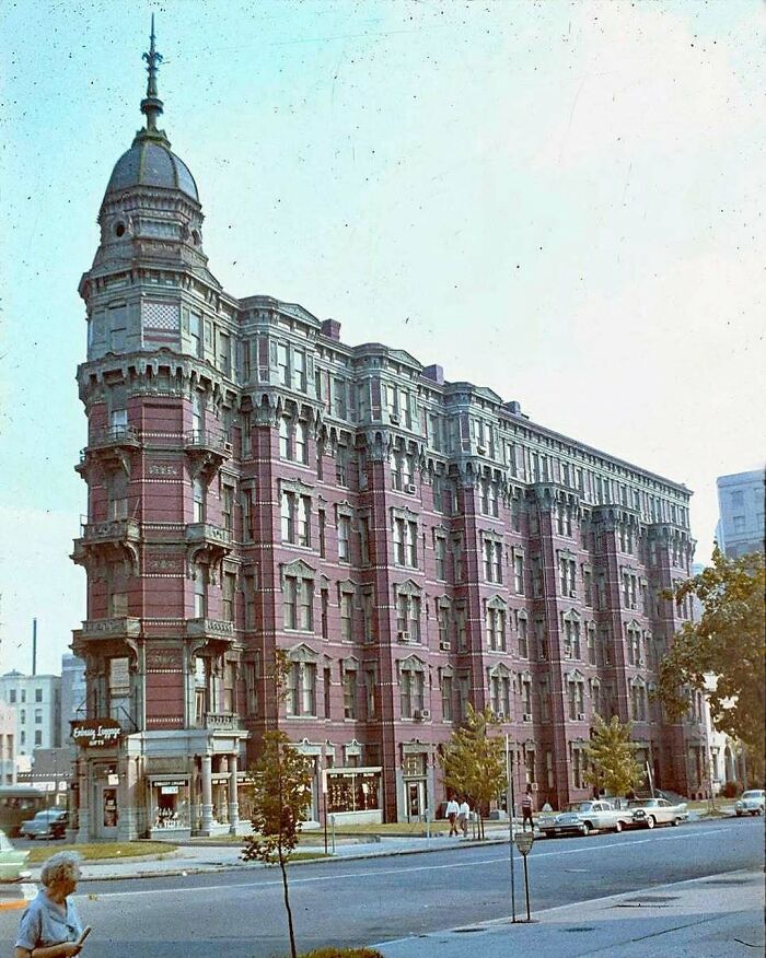 Historic American architecture with ornate details on a multi-story red brick building, cars, and pedestrians nearby.