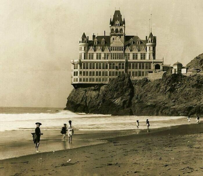 Historic architecture on a cliff by the seaside, people walking along the beach below.