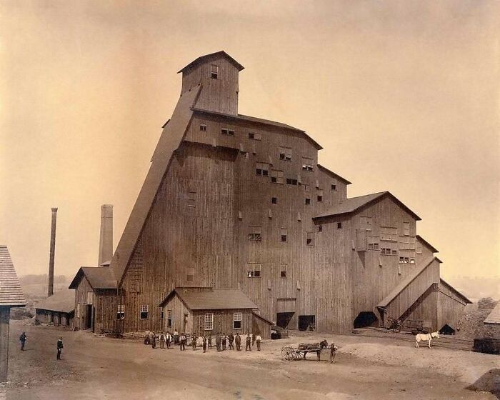 Historic American architecture with people and horses in front of a large wooden industrial building.