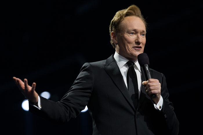 Man in a suit holding a microphone, delivering a monologue on stage with a spotlight backdrop, relating to controversial jokes at Oscars. Man in a suit holding a microphone, delivering a monologue on stage with a spotlight backdrop, relating to controversial jokes at Oscars.