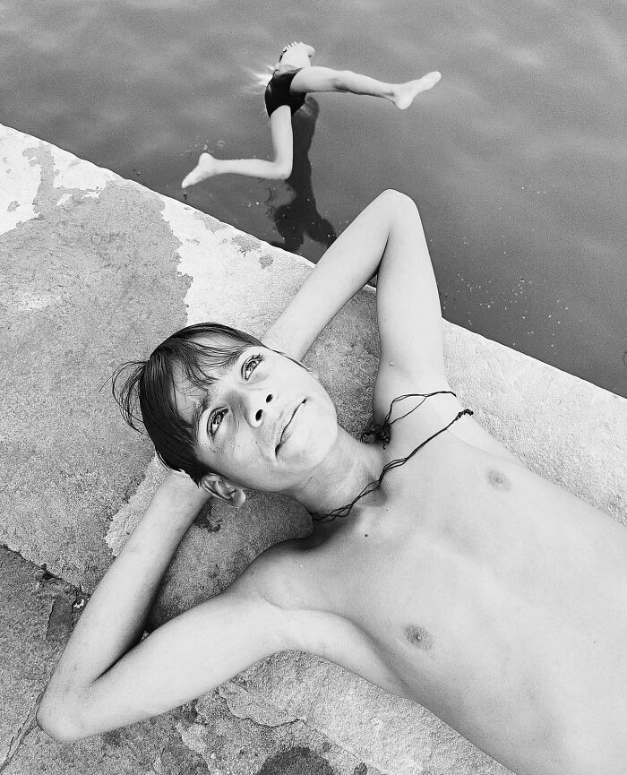 Boy rests on a concrete edge while another leaps into water in a split-second black and white photo.