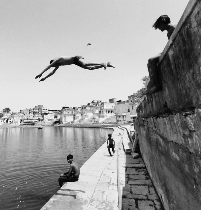 Child mid-air in split-second leap over river, B&W photography, others watch from old city banks.