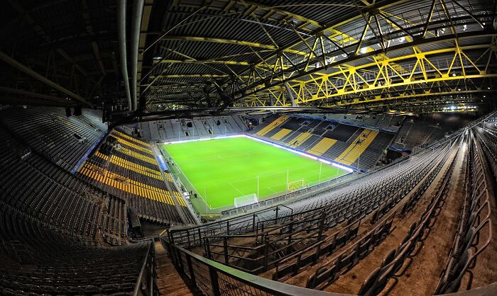 Interior view of a large cathedral of soccer stadium with empty seats and a brightly lit green pitch.