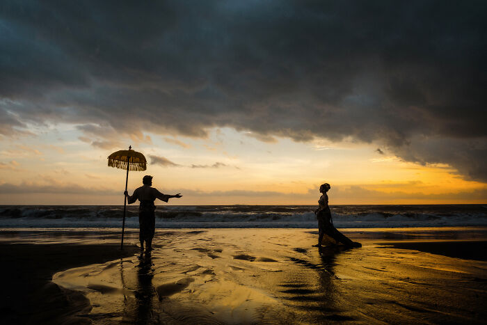 Sunset beach engagement photo with a silhouetted couple, one holding an umbrella, under dramatic clouds.