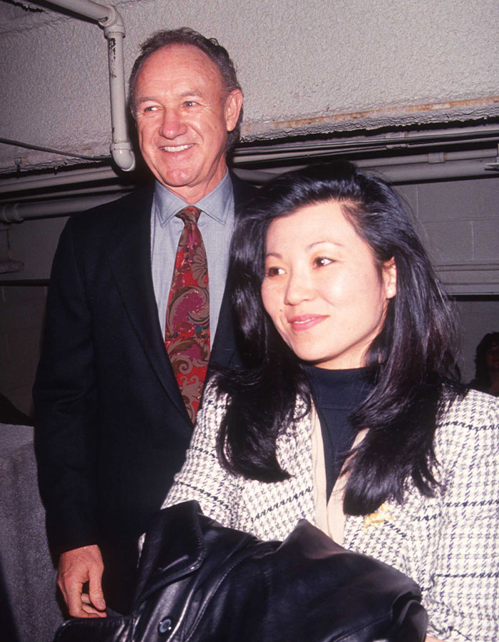 Elderly man in a suit smiling, standing next to a woman in a checkered jacket; focus on Gene Hackman topic. Elderly man in a suit smiling, standing next to a woman in a checkered jacket; focus on Gene Hackman topic.