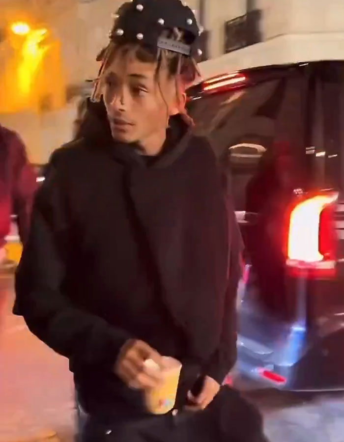 Jaden Smith with unique style, holding a drink near a car at night. Jaden Smith with unique style, holding a drink near a car at night.