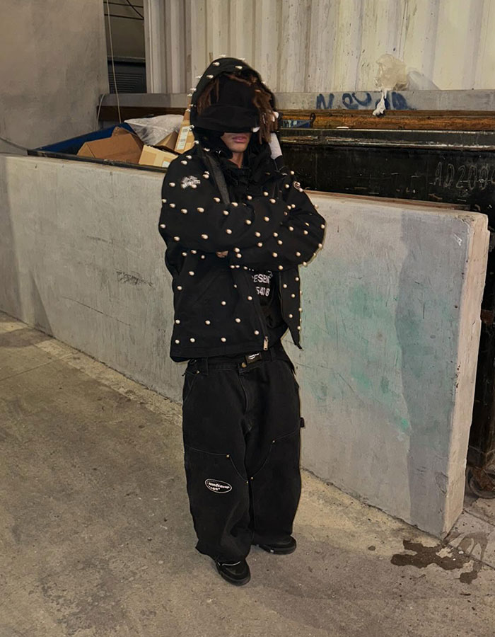 Young man in a dark outfit with hood up, standing in an industrial setting, sparking concern with his appearance. Young man in a dark outfit with hood up, standing in an industrial setting, sparking concern with his appearance.